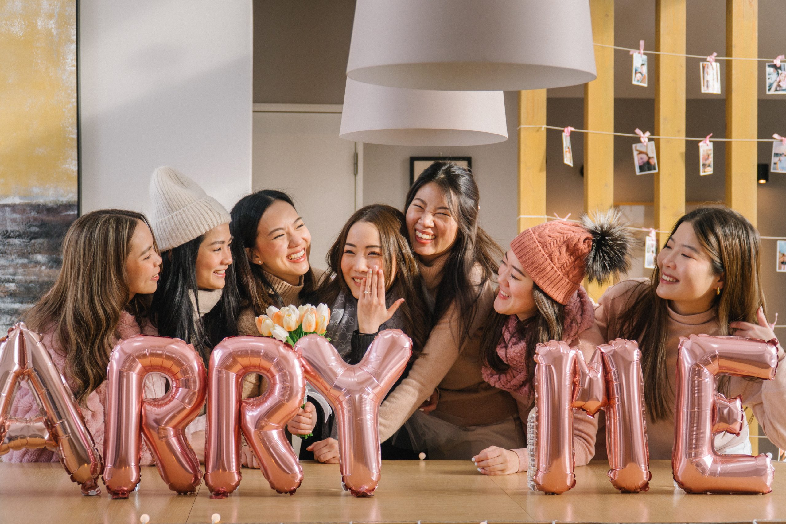 Friends celebrating a proposal beside Marry Me balloons